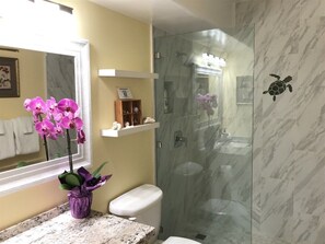 Ein Badezimmer mit Duschkabine aus Glas, Toilette, Wand in Marmoroptik und einer Vase mit Blumen auf dem Waschbecken.