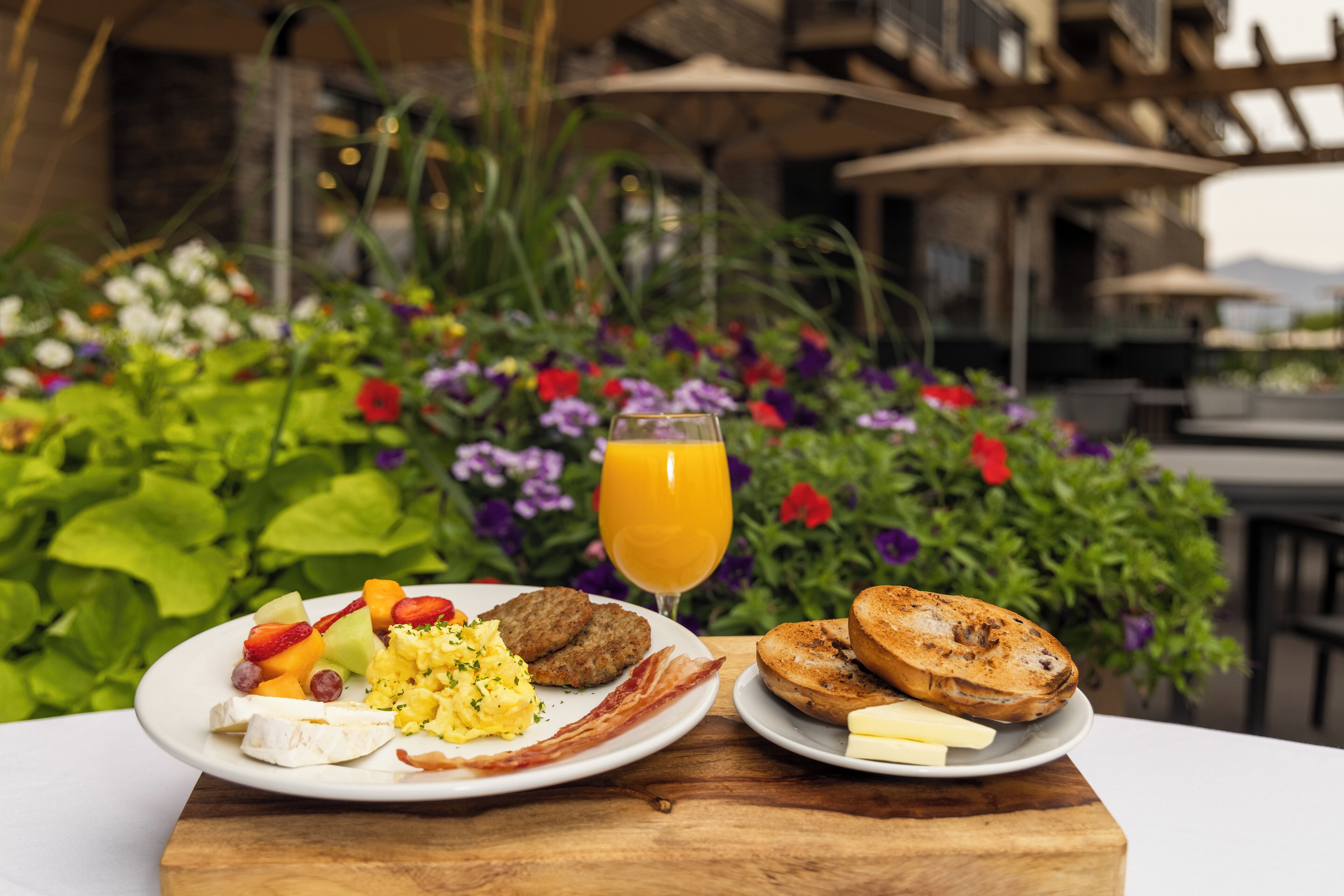 A breakfast table with plates of food, a glass of orange juice, and a view of a garden with flowers and plants.