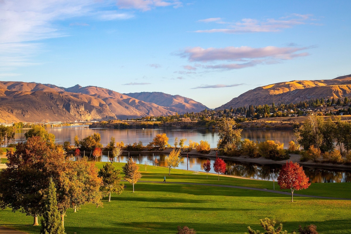A serene lakeside landscape with mountains in the background, a clear sky, and a well-maintained grassy area with trees.