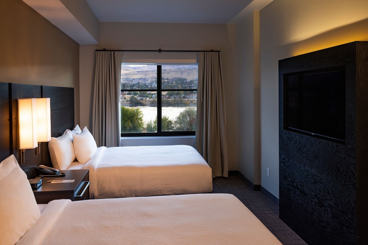 A hotel room with two beds, a large window with curtains, a flat-screen TV, and a bedside table with a lamp and phone.