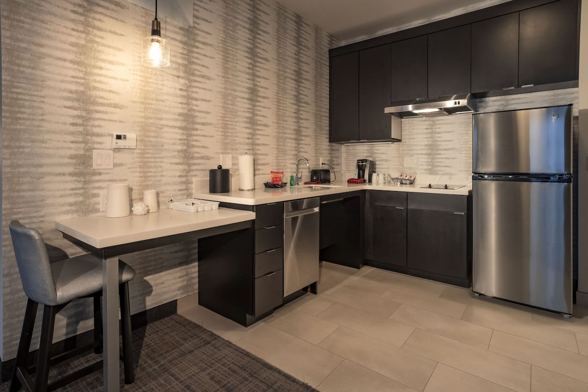 A modern kitchen with dark cabinets, a stainless steel refrigerator, a built-in dishwasher, and a light fixture.