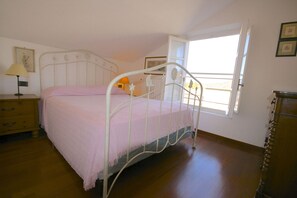 A bedroom with a metal bed frame, a wooden dresser, a bedside table with a lamp, and a window with a view of the outside.