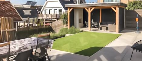 A patio area with a table and chairs, a green lawn, and a covered seating area with a view of neighboring houses.
