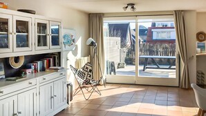 Een lichte woonkamer met een schuifdeur naar buiten, een wit kastje met glazen deuren, een vloerlamp en een kleine tafel.