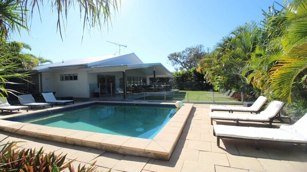 A swimming pool with lounge chairs, a covered patio, and tropical vegetation.