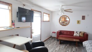 Una sala de estar con un sofá rojo, una mesa blanca, un televisor, un ventilador de techo y una ventana.