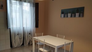 A dining area with a white table and chairs, a painting of cacti on the wall, and a window with sheer curtains. - Spacious apartment near the beaches and at the foot of Mount Etna (Mascali)