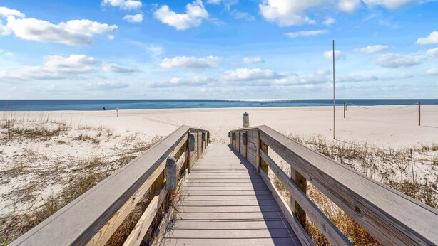 Ein hölzerner Steg führt zu einem Sandstrand unter blauem Himmel.