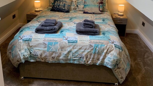 Ein Schlafzimmer mit schrägverdachter Decke, einem Bett mit Bezug im Küstenstil, zwei Nachttischlampen und einem Oberlicht.