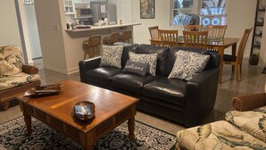 A living room with a black leather sofa, patterned cushions, a wooden coffee table, and a patterned rug. There is a dining area with wooden chairs and a table, and a kitchen in the background. - Beautiful home in Kailua-Kona with easy access to all the Island. (KAILUA KONA)