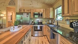 Cocina de cabaña de madera con refrigerador, horno y microondas de acero inoxidable, armarios de madera, encimera de granito y ventana con vistas a la vegetación.