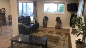 A living room with a black leather sofa, a coffee table, and a patterned rug. There are potted plants and a television mounted on the wall. - Spacious one bedroom suite (Campbell River)
