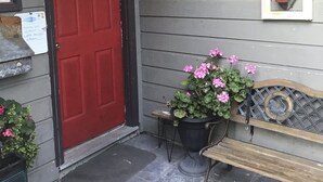 A front porch with a red door, a bench, a potted plant with pink flowers, and a wall-mounted decoration. - Spacious one bedroom suite (Campbell River)
