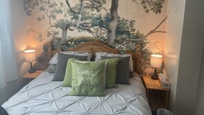 A bedroom with a large tree mural on the wall, a bed with a white quilted bedspread, and two bedside lamps. - Spacious one bedroom suite (Campbell River)