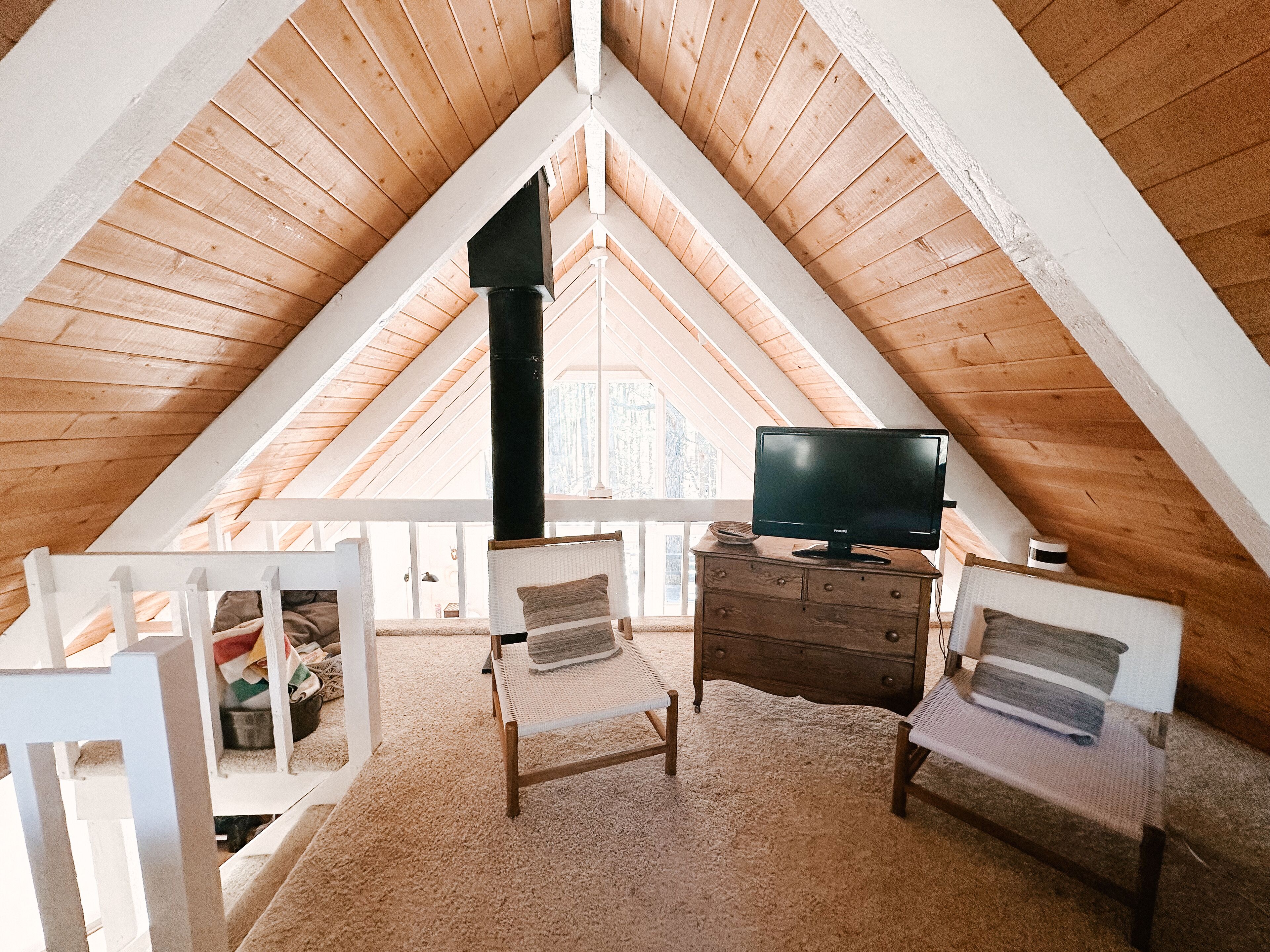 A cozy attic bedroom with a sloped ceiling, a television on a dresser, two chairs, and a large window offering a view of the outdoors.