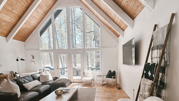 Ein geräumiges Loft mit hoher Decke, Holzbalken, einem großen Fenster mit Blick auf Bäume, einer gemütlichen Sitzecke mit Sofa und Sesseln und einem an der Wand befestigten Fernseher.