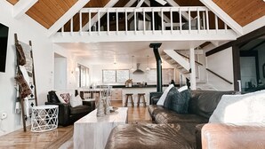 Ein geräumiger Wohnbereich mit hoher Decke, Holzbalken, einem großen Ledersofa und einer Küche im Hintergrund.