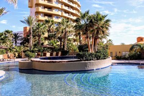 A resort pool area with palm trees, lounge chairs, and a multi-story building in the background.