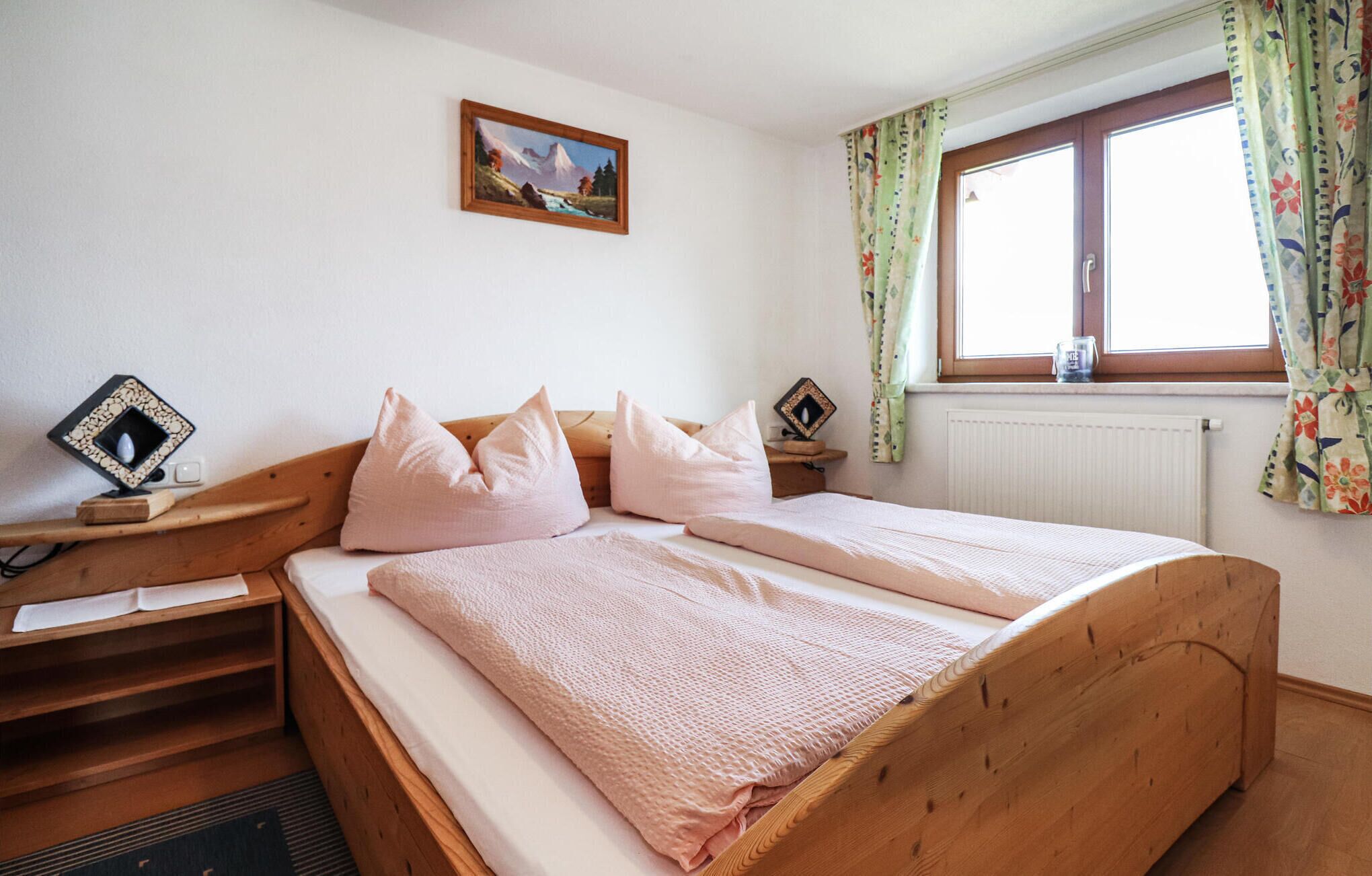 A bedroom with a wooden bed, a small bedside table, a nightstand, a framed picture on the wall, and a window with curtains.