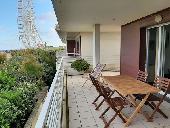 A balcony with a wooden table and chairs, a potted plant, and a view of a Ferris wheel.