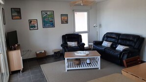 Sala de estar con un sofá de cuero negro, una mesa de centro de madera y un televisor sobre un mueble bajo. Se observan cuadros enmarcados en la pared y una ventana con luz natural.