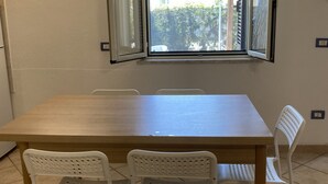 Ein rechteckiger Esstisch mit vier weißen Stühlen, ein Fenster mit geöffneten Rollläden und eine weiße Wand mit Steckdose.