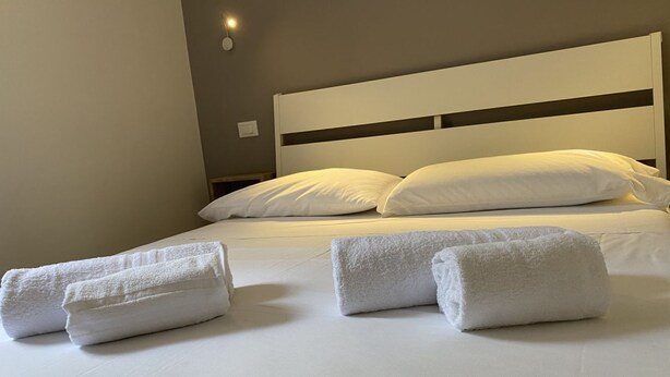 Un letto rifatto con lenzuola e cuscini bianchi, un comodino con una lampada e una luce a parete.