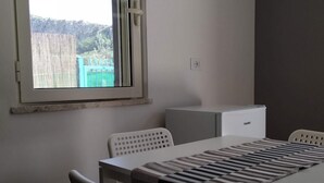 Una stanza piccola e moderna con un tavolo e sedie, una finestra che si affaccia su una recinzione e degli alberi, e un piccolo elettrodomestico bianco a parete.