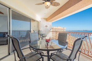 A balcony with a glass table and chairs, a ceiling fan, and a view of the sea and buildings.