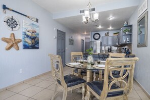 A dining area with a round glass table, wooden chairs with cushioned seats, a chandelier, and nautical-themed wall decorations.