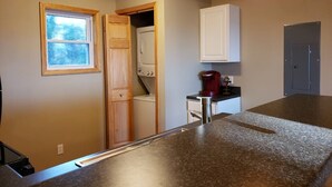 A kitchen with a granite countertop, a built-in washer and dryer, a coffee maker, and a window with a view of trees. - Private beach location (Oscoda)