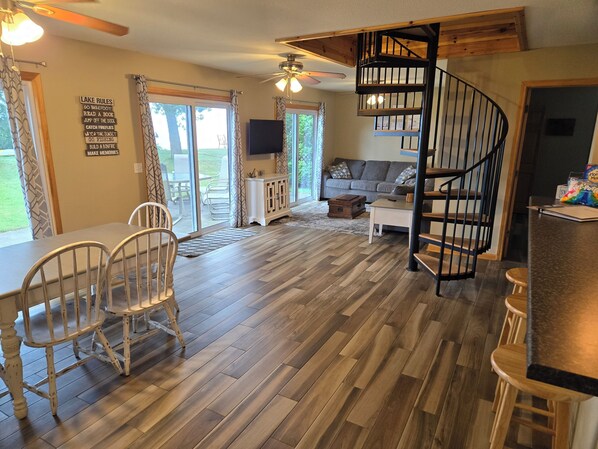 A spacious living area with a wooden floor, a dining table with chairs, a sofa, a TV, and a spiral staircase. - Private beach location (Oscoda)