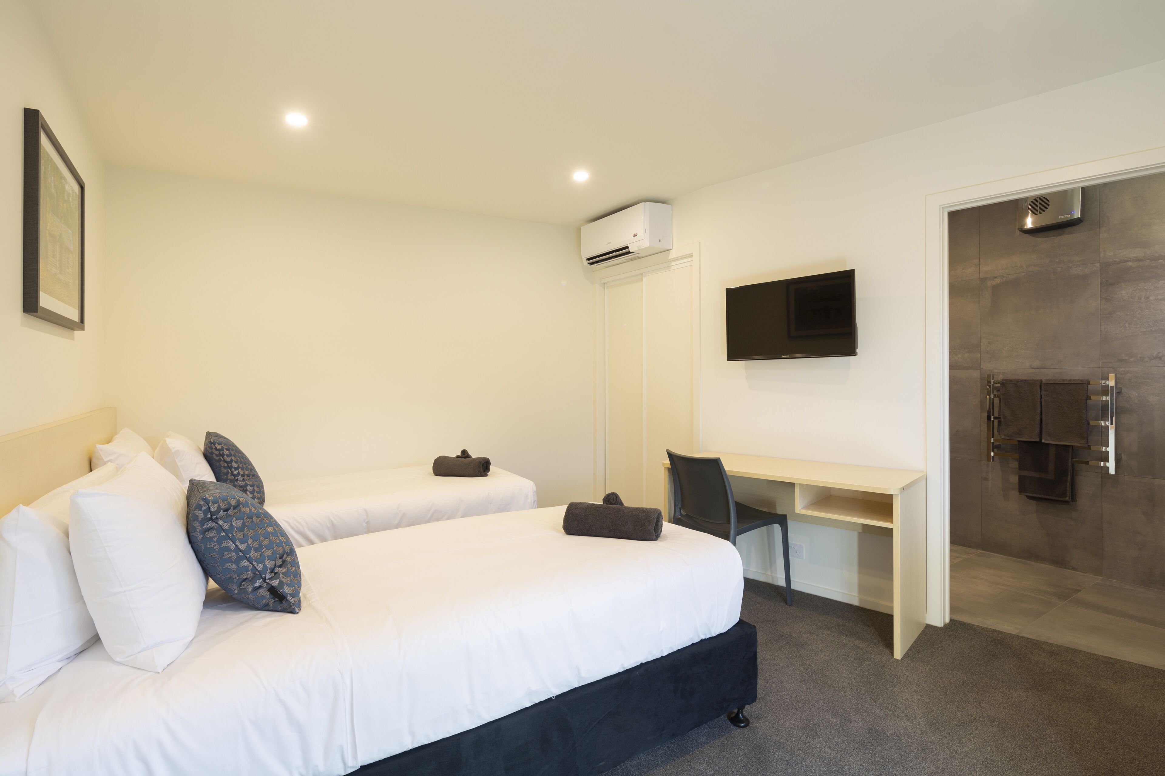 A hotel room with two beds, a desk, a flat-screen TV, and an air conditioning unit.