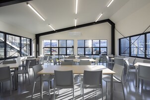 Um espaço amplo e interno com várias mesas e cadeiras, grandes janelas com vista para edifícios e montanhas, e um design moderno no teto.