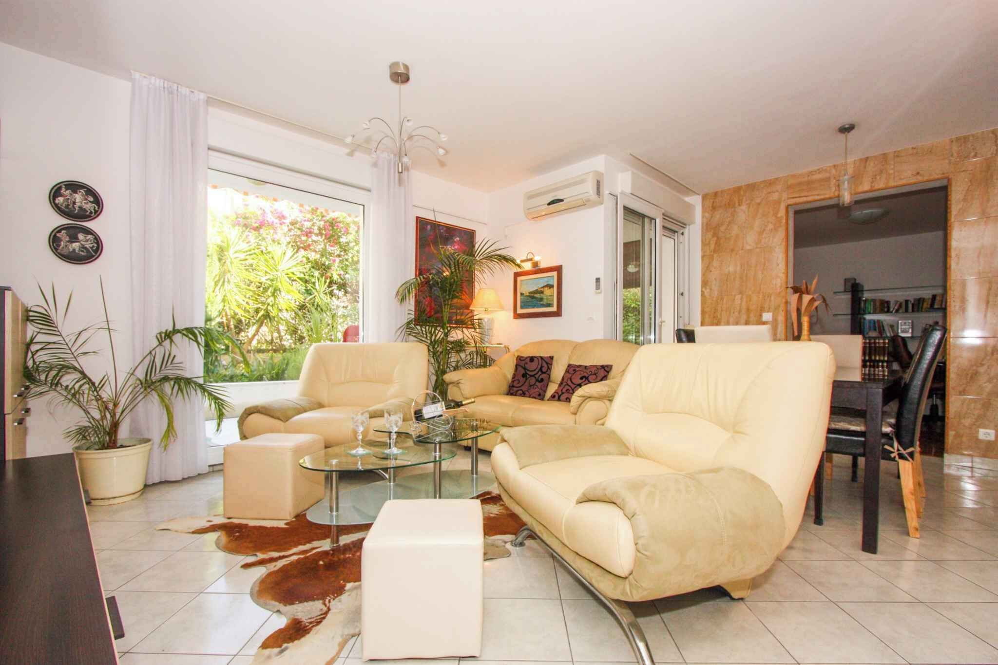 A living room with a beige sofa, a glass coffee table, and a dining area with a wooden table and chairs. There are potted plants and a painting on the walls.