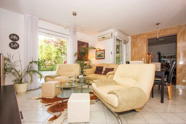 A living room with a beige sofa, a glass coffee table, and a dining area with a wooden table and chairs. There are potted plants and a painting on the walls.