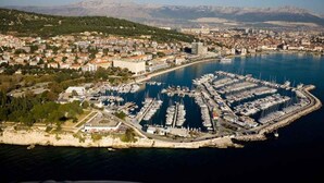 Vista aérea de uma vila costeira com um porto de iates, edifícios e montanhas ao fundo.