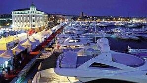 Uma marina com vários iates atracados, um edifício grande com cúpula e tendas ao longo da orla.