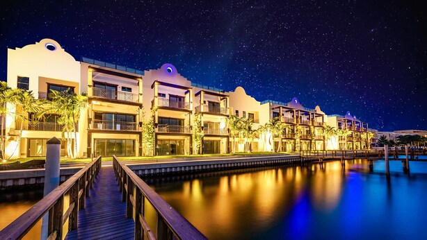 A waterfront hotel with multiple balconies, a wooden boardwalk, and a calm water body reflecting the night sky.