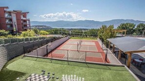 A tennis court with a surrounding fence, a covered seating area, and a view of mountains in the background. - Pools open May long weekend!  Holiday Time @ West Kelowna's hidden gem!  2BR (West Kelowna)