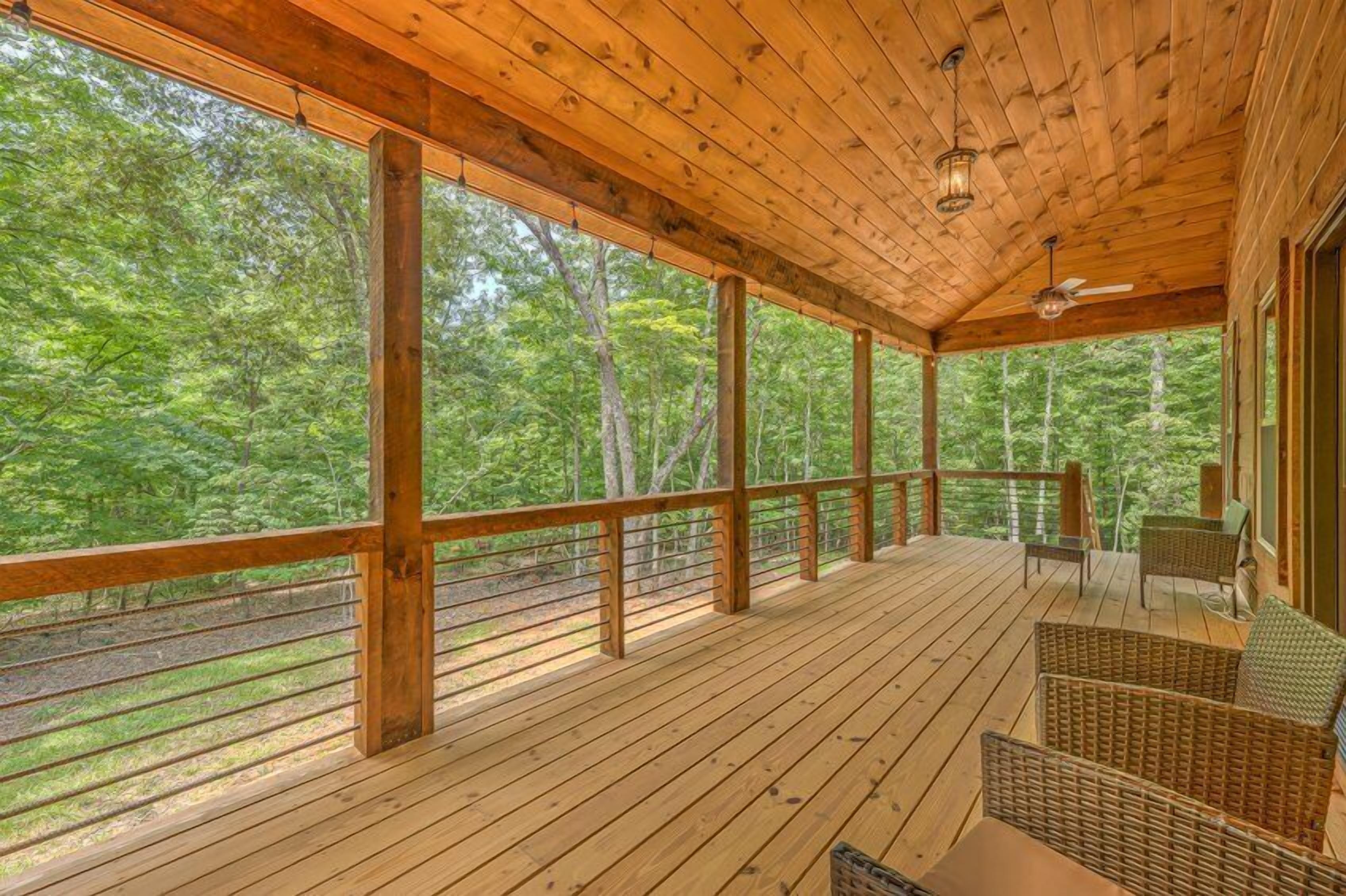 Una terraza de madera con mobiliario de mimbre, ventanas amplias y vista a un bosque.