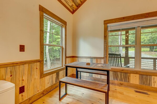 Una habitación con paredes y suelos de madera, ventanas grandes con vistas a árboles, un banco de madera y una pequeña mesa.
