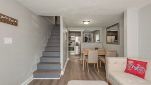 Una cocina moderna con electrodomésticos de acero inoxidable, un comedor con sillas de madera y una escalera que sube a otro nivel.