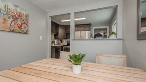 Un comedor con una mesa de madera, sillas y una planta en maceta. Se ve una cocina al fondo, con armarios y electrodomésticos.