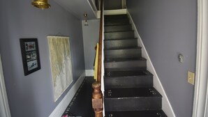 A staircase with black steps and a wooden handrail, leading to a landing with a light fixture and a framed picture on the wall. - Relax at the historic Austin House (Addison)