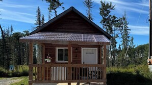 Una cabaña de madera pequeña con techo metálico, una puerta principal y un porche con un banco.