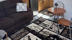 Una sala de estar acogedora con un sofá marrón, un tapete estampado, una mesita redonda con una silla y armarios de cocina de madera.