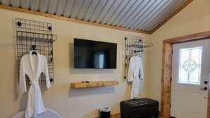 A room with wooden flooring, a television mounted on the wall, a wooden shelf, a white door with a window, and two white robes hanging on wire racks. - Barn Loft at Private Retreat - Horses Welcome (Boyd)