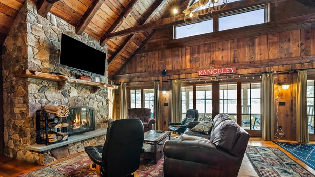 A cozy living room with a stone fireplace, leather furniture, and a large window overlooking the outside. - Bishop | Amazing location on Rangeley Lake and close to downtown (Rangeley)