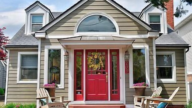 Una casa de dos pisos con una puerta principal roja, un porche con dos sillas y una corona en la puerta.